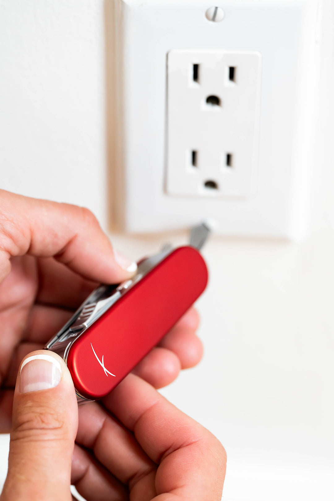 A person using the screwdriver of a Sizzle Scout USB rechargeable lighter/survival device in red color to tighten a screw of an electrical outlet cover